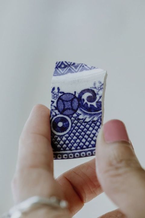 Porcelain. A person holds a roughly rectangular fragment of white porcelain painted with blue patterns and leafy designs between a thumb and forefinger. The thumbnail is painted pink.