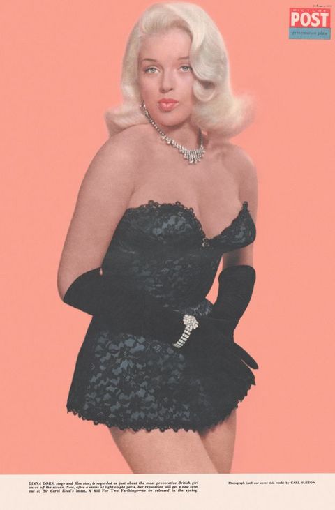 Shown from the thighs up, a pale-skinned woman with blond hair stands in front of a vivid peach background. She wears a strapless black bustier and her short skirt brushes the top of her thighs. Black gloves reach to her elbows, and she wears a diamond necklace and bracelet. Her pink lips are pursed.