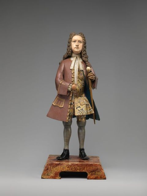 Sculpture. This free-standing clay statuette shows a pale-skinned man wearing a long, brown wig over a mauve-pink coat edged with gold. One side of the coat is held back with a walking stick to show a gold patterned vest beneath. He wears grey stocking and stands squarely in his black shoes.