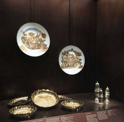 Installation. Two porcelain plates and eight pieces of metalwork are displayed in a case set into a wall. The plates hang on the wall over five gold-coloured dishes and three silver casters. Explanatory text can be seen (but not read) below the display.