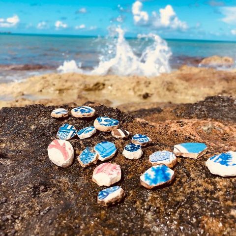 Porcelain. Seventeen roughly round pieces of broken ceramic are laid out on a rock in front of an ocean. The fragments are mostly blue and white but two have red on white and one has brown. A breaking wave splashes in the distance, which is out of focus.