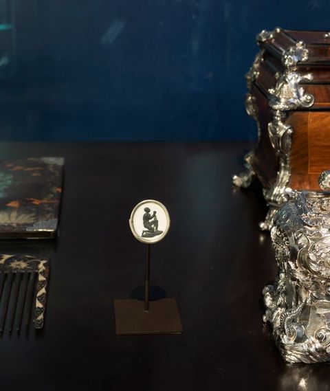 Installation. A white oval with a black person almost in silhouette id displayed on a stand next to a comb and a silver object. On the medallion, a man kneels and lifts his hands. Chains hang from shackles around his wrists, and he looks up.