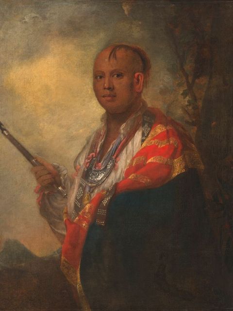 Painting. Shown from the waist up, a medium-skinned man’s body is angled to our left, but he turns to look at us from the corners of his dark eyes. A lock of straight hair falls over his forehead, which is bald over the crown. He wears a white shirt, a crescent-shaped metal gorget low on his chest, and a navy-blue and crimson-red cloak edged with gold.