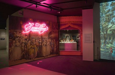Installation. A floating wall is covered with an enlarged print showing women wearing long gowns and men wearing hats and suits. The title of the exhibition is in pink neon, illuminating the otherwise dim space. Pink-glowing footsteps seem to emerge from the print and turn the corner, leading us into the exhibition.